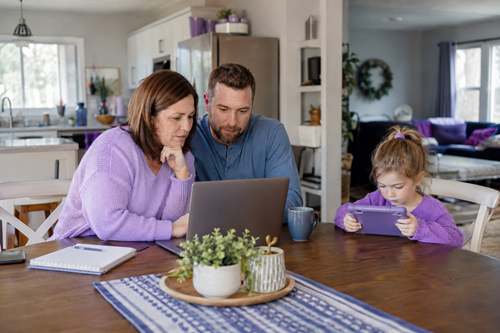A couple reviews internet plan options on a laptop at the kitchen table while a child watches a tablet nearby.